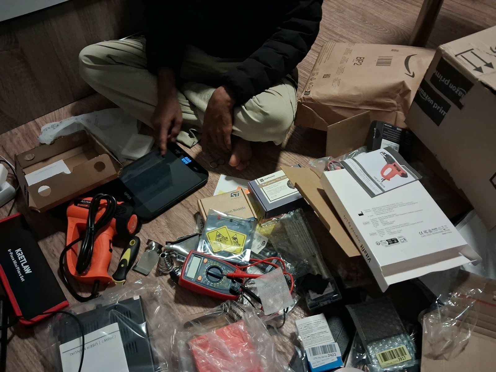 Parts and tools spread across a wooden floor: a multimeter, a heat gun, pliers, a scale and the ReSpeaker microphone array box, with open delivery boxes behind.