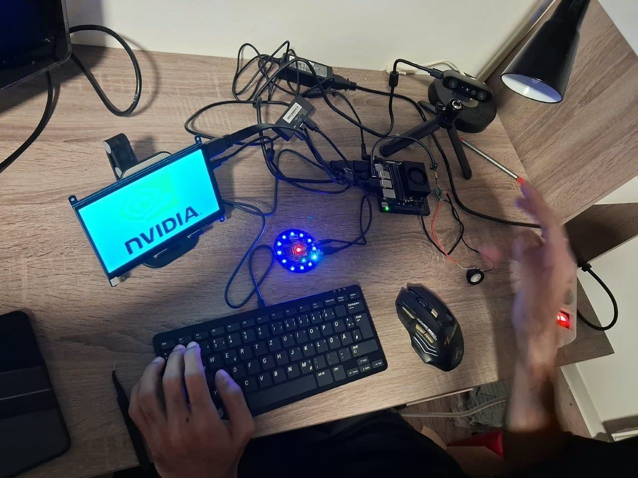 A desk seen from above: the 7 inch screen showing the NVIDIA logo, the Jetson with its fan, the microphone ring lit blue and the depth camera on a small tripod.