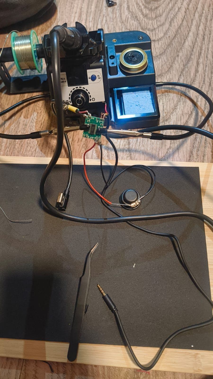 A small green amplifier board soldered to a round speaker and an audio cable, next to a soldering station and tweezers on a black mat.